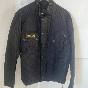 Svart quiltad jacka från Barbour - Säljer en snygg svart quiltad jacka från Barbour International. Design med dragkedja och knappar framtill, samt en broderad logga på bröstet. Perfekt för höst och vår! 🖤