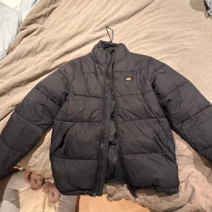 Svart pufferjacka från Dickies - Säljer en svart pufferjacka från Dickies, nypris 1100kr. Jackan är i bra skick, dock har den en lagning på ena armen, därav säljs den för 450kr. Jackan har en dragkedja framtill och är perfekt för kyliga dagar. Den är vadderad och har en normal passform med två ytterfickor och två innerfickor. Perfekt för höst och vinter!