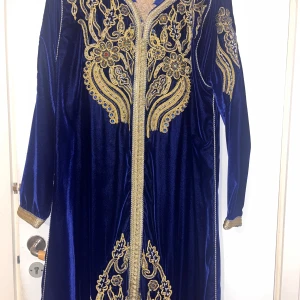 Blå sammet klänning med guldbroderi - Elegant långärmad klänning i mörkblå sammet med detaljerat guldbroderi. Perfekt för festliga tillfällen med sin lyxiga känsla och vackra mönster. Klänningen har en normal passform och är i mycket bra skick.