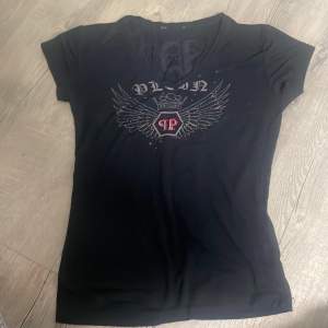 Säljer en svart t-shirt från Philipp Plein med coola vingdetaljer, paljettglitter detaljer och slitsar på ryggen. Den har en v-ringad hals och korta ärmar, samt jätte mjukt material