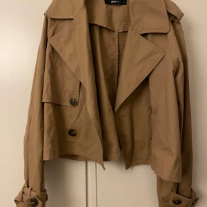 Beige trenchcoat från Gina Tricot - Säljer en snygg beige trenchcoat från Gina Tricot i storlek S. Jackan har en klassisk design med stora knappar och bälte vid ärmsluten. Perfekt för höst och vår, och ger en stilren look till vilken outfit som helst.