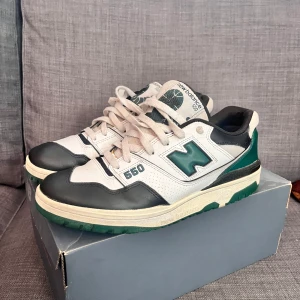 New Balance 550 sneakers - Säljer ett par New Balance 550 sneakers i vitt, svart och grönt. De är i relativt bra skick och har en snygg retrostil. Perfekta för både vardag och träning. Storlek 45. De har en liten orange fläck under snöret, men syns inte vid användning. Ge dom en tvätt så har du ett par riktigt fina skor.