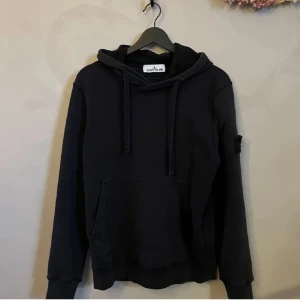 Svart hoodie från Stone Island - Säljer en snygg svart hoodie från Stone Island i bra skick. Den har en klassisk design med känguruficka och justerbar huva. Perfekt för både vardag och chill. Märkesloggan sitter på ärmen för en stilren look. Passar perfekt till jeans eller joggers! Har ett litet hål i armen men inget som syns eller märks! Pris ej hugget i sten!! Kom privat för mer info🙂      MVH Emil