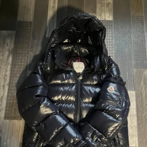 Moncler maya  - Säljer en snygg svart dunjacka från Moncler i mycket bra skick. Jackan har en glansig finish och är perfekt för kalla vinterdagar. Den har en dragkedja framtill och en huva för extra värme. Moncler-loggan är broderad på ärmen. Perfekt för dig som vill hålla stilen i vinter! ❄️
