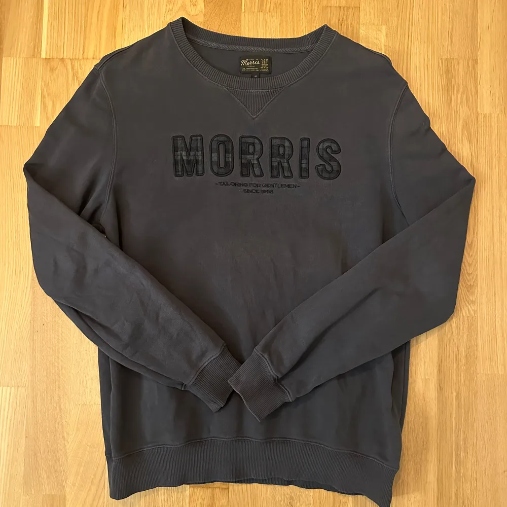 En grå sweatshirt med en broderad logotyp på framsidan. Logotypen är i svart och vitt med en rutig design. På baksidan av tröjan finns ingen text eller design. Tröjan är hel och ren men lite smått blekt runt kragen. Se bilder. Storlek M. Tröjor & Koftor.