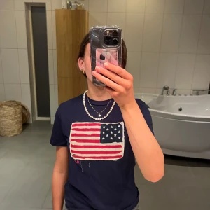Mörkblå Ralph Lauren t-shirt - Säljer en mörkblå t-shirt med en stor amerikansk flagga på framsidan. T-shirten är kortärmad och har en normal passform. Perfekt för en avslappnad stil eller sommarens fester. Passar bra till jeans eller shorts! Säljes då den inte passar längre då den är i storlek 152-164