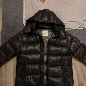 Säljer en snygg svart dunjacka från Moncler i mycket bra skick. Jackan har en huva och dragkedja framtill, perfekt för kalla vinterdagar. Den är vadderad och har Monclers logga på ärmen. Insidan har en cool serietidningsdesign. Passar både till vardags och lite finare tillfällen.