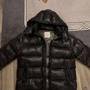 Moncler maya jacka - Säljer en snygg svart dunjacka från Moncler i mycket bra skick. Jackan har en huva och dragkedja framtill, perfekt för kalla vinterdagar. Den är vadderad och har Monclers logga på ärmen. Insidan har en cool serietidningsdesign. Passar både till vardags och lite finare tillfällen.