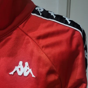 Röd träningsjacka från Kappa - Säljer en snygg röd träningsjacka från Kappa. Den har en dragkedja framtill och långa ärmar med det klassiska Kappa-mönstret längs axlarna och ärmarna. Perfekt för sportiga aktiviteter eller som en stilren vardagsjacka. Passar bra till både träning och chill.