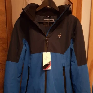 Blå och svart vindjacka - Säljer en snygg blå och svart vindjacka från 2117 of Sweden. Jackan har en dragkedja framtill och praktiska fickor med dragkedjor. lappen kommer ej,  Perfekt för blåsiga dagar och utomhusaktiviteter. Den har en normal passform och är långärmad. Passar både till vardags och lättare friluftsaktiviteter.