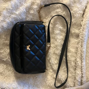 Svart quiltad axelväska med gulddetalj - Snygg svart axelväska i quiltat mönster med en söt guldfärgad fjäril på framsidan. Väskan har en lång axelrem med kedjedetaljer och är perfekt för både vardag och fest. Den är i mycket bra skick och redo att användas direkt!