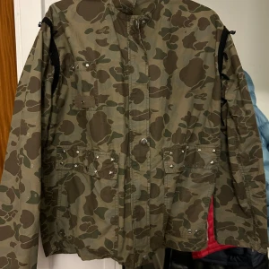 Camouflage jacka med nitar - Säljer en cool camouflage jacka med nitar på fickorna. Jackan har långa ärmar och flera praktiska fickor. Perfekt för höst och vår, och ger en edgy look till din outfit. Passar bra för casual tillfällen eller när du vill sticka ut lite extra.