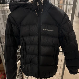 Svart dunjacka från Peak Performance - Säljer en svart dunjacka från Peak Performance i mycket bra skick. Jackan har en dragkedja framtill och en praktisk huva. Perfekt för kalla vinterdagar. Den är både stilren och funktionell med flera fickor. Nypris är runt 3000 kr och säljer denna för 2000. 