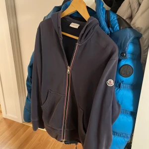 Mörkblå hoodie från Moncler - Säljer en snygg mörkblå hoodie från Moncler i mycket bra skick. Den har en dragkedja framtill och en klassisk Moncler-logga på ärmen. Perfekt för höst och vår, och den är superbekväm med sin mjuka bomullskvalitet. Passar perfekt för en casual look eller när du bara vill mysa hemma. Skriv för mer bilder och priset kan alltid diskuteras!