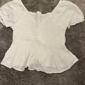 Vit blus med volangdetaljer - Säljer en söt vit blus med volangdetaljer. Den har en smockad design som ger en fin passform och puffiga korta ärmar. Perfekt för sommardagar eller en avslappnad vårlook. Blusen är i mycket bra skick och redo för nya äventyr! 