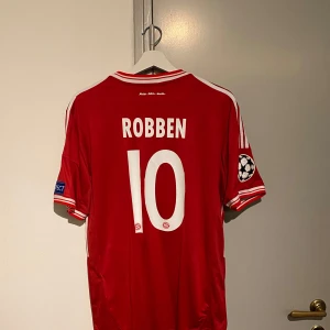 Röd Bayern München fotbollströja med Robben 10 - Säljer en röd Bayern München fotbollströja från Adidas med Robben och nummer 10 på ryggen. Tröjan är kortärmad och har Champions League-märken på ärmarna. Framsidan har Adidas-logga och T-Mobile sponsortryck. Perfekt för samlare eller fans av Bayern München!