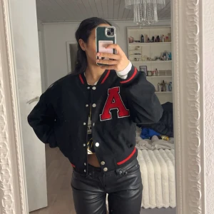 Svart bomberjacka med röd bokstav - Säljer en snygg svart bomberjacka med en stor röd bokstav 'A' på bröstet. Jackan har knappar framtill och röda detaljer vid krage, ärmslut och nederkant. Perfekt för en cool och avslappnad stil. Passar bra till både vardag och fest!