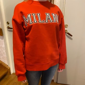Röd sweatshirt - En röd sweatshirt med texten 'MILANO' i vitt och svart på bröstet. Tröjan har en avslappnad passform och långa ärmar, perfekt för en casual look. Den är i bra skick och passar perfekt till hösten och vintern.