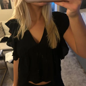 Svart blus med volangdetaljer - Säljer en snygg svart blus med volangdetaljer. Den har korta ärmar och en luftig passform, perfekt för både vardag och fest. Blusen är i mycket bra skick 💕