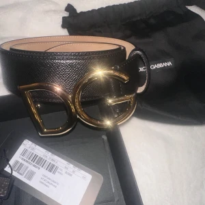 Svart skärp från Dolce & Gabbana - Säljer ett snyggt svart skärp från Dolce & Gabbana i skinn med en stor, glittrig DG-spänne i guld. Perfekt för att lyfta vilken outfit som helst! Kommer i originalförpackning och är i nytt skick. Passar perfekt till fest eller en stilren vardagslook. 90cm långt. Obs. Något små slitage på guldfärgen