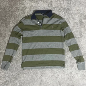 Randig pikétröja från GANT - Säljer en snygg randig pikétröja från GANT i grönt och grått. Tröjan har långa ärmar och en klassisk krage med knappar. Perfekt för en avslappnad men stilren look. Passar bra till både jeans och chinos!