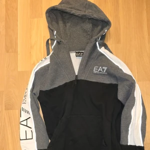 Grå och svart hoodie från Emporio Armani Stl. S - Säljer en snygg grå och svart hoodie från Emporio Armani. Den har en dragkedja framtill och en stor luva. Ärmarna är dekorerade med vita detaljer och loggan EA7 tryckt på bröstet och ärmen. Perfekt för en casual look eller träning. Hör av dig vid intresse! 