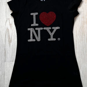 Svart t-shirt med I ❤️ NY-design - Snygg svart t-shirt med det ikoniska 'I ❤️ NY'-trycket i glittriga stenar. Perfekt för en casual look eller en utekväll. T-shirten är kortärmad och har en normal passform. Ett måste för alla som älskar New York!