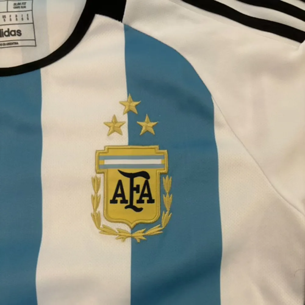 Säljer en snygg Argentina fotbollströja från Adidas i storlek S. Tröjan har de klassiska blå och vita ränderna med AFA-emblemet på bröstet. Den är kortärmad och har svarta detaljer vid krage och ärmslut. Perfekt för fotbollsfans!. T-shirts.