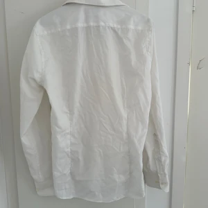 Riley ”Erik Regular Skjorta Twill Vit” -  En vit Riley-skjorta är en klassisk och stilren skjorta i mjukt material. Den har krage, knappar framtill och långa ärmar, vilket gör den perfekt för både formella och vardagliga tillfällen.