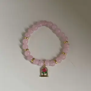 Säljer ett superfint armband med ljusrosa pärlor och guldfärgade detaljer. Armbandet har en söt charm med en blomma i rosa och grönt. Perfekt för att ge en färgklick till din outfit!