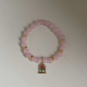 Rosa armband med gulddetaljer och blomcharm - Säljer ett superfint armband med ljusrosa pärlor och guldfärgade detaljer. Armbandet har en söt charm med en blomma i rosa och grönt. Perfekt för att ge en färgklick till din outfit!