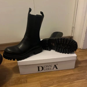 Svarta boots från Donn A Girl - Säljer ett par svarta boots från Donn A Girl i storlek 38. De är gjorda i syntetmaterial och har en rejäl, grovmönstrad sula som ger bra grepp. Bootsens stilrena design gör dem perfekta för både höst och vinter. De har en dragkedja på sidan för enkel på- och avtagning.