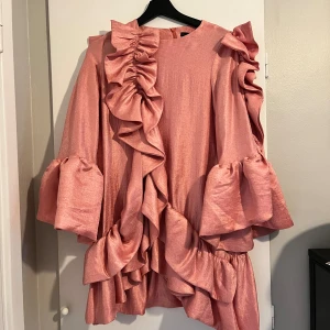 Rosa klänning med volanger från Mummymoon - Säljer en superfin rosa klänning från Mummymoon. OBS storlek 12 i barnkläder. Den har en lös passform med volanger som ger en härlig volym. Klänningen är långärmad och har en dragkedja i ryggen. Perfekt för festliga tillfällen! 💖 passar XS