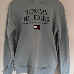 Ljusblå hoodie från Tommy Hilfiger - Säljer en snygg ljusblå hoodie från Tommy Hilfiger. Den har ett stort tryck med märkesnamnet på framsidan och är superbekväm. Perfekt för en avslappnad stil. Den är i bra skick och har ribbade muddar vid ärmslut och nederkant. Djur- och rökfritt hem