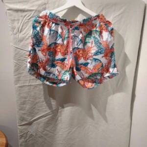 Färgglada blommiga shorts - Säljer ett par superhärliga shorts med ett färgglatt blommönster. De har en elastisk midja med rosa snörning för justerbar passform. Perfekta för sommardagar på stranden eller en avslappnad dag i parken. Materialet känns lätt och luftigt. Aldrig använda