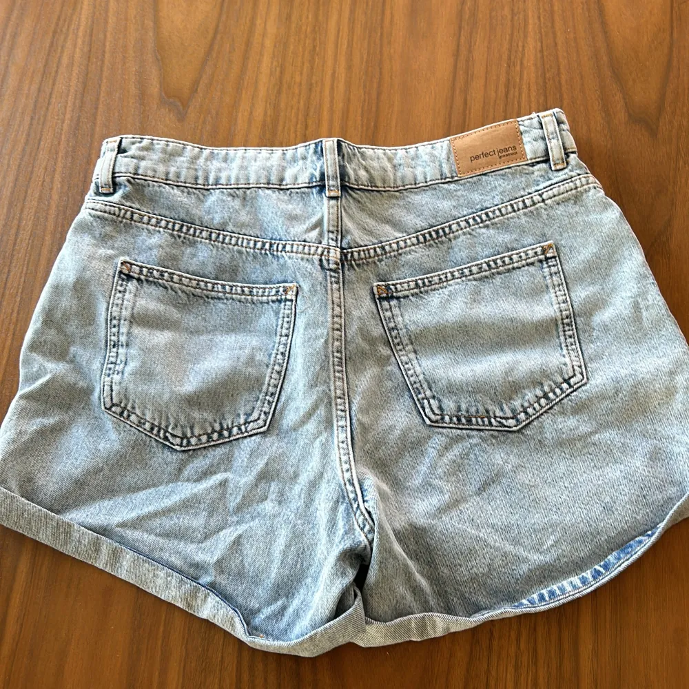 Säljer ett par klassiska jeansshorts från Perfect Jeans i storlek 42. De är ljusblå och har en avslappnad passform med uppvikta ben. Shortsen har fem fickor och stängs med en knapp och dragkedja. Perfekta för sommardagar!. Shorts.