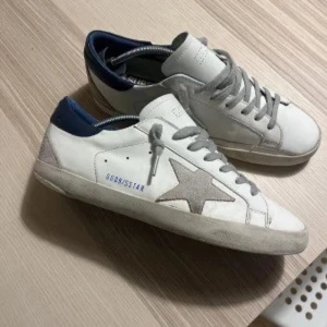 Vita sneakers med grå stjärna - Snygga vita sneakers från Golden Goose med en grå stjärna på sidan och grå snörning. Skorna har en mörkblå häl och en vintage-inspirerad look. Perfekta för en avslappnad stil.