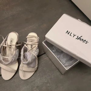 Klackar från NLY Shoes - Eleganta beige klackar med klack från NLY Shoes. Skorna har en stilren design med knytning runt ankeln och dekorativa stenar på remmen. Helt nya. 