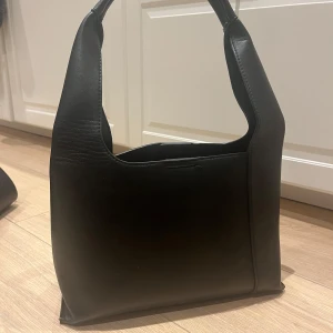 Svart axelväska i skinn - Väskan har en enkel och elegant form med en praktisk öppning och stängning. Perfekt för att bära dina dagliga nödvändigheter med stil.