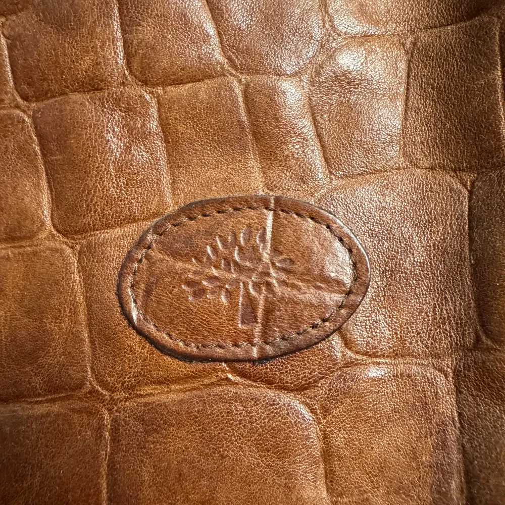 Snygg brun axelväska i skinn med krokodilpräglat mönster. Väskan har ett emblem på framsidan och en metallknapp med ett trädmotiv. Insidan är fodrad med ett rutigt tyg. Perfekt för en stilren look. ÄKTA. . Laukut & Käsilaukut.