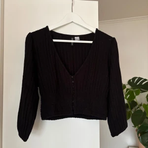 Svart ribbad kofta - Snygg svart ribbad kofta med v-ringning och knappar framtill. Den har trekvartsärmar och en croppad passform, perfekt för lager på lager-stil. En stilren och mångsidig topp för olika tillfällen.