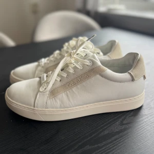 Vita sneakers från Calvin Klein - Snygga vita sneakers från Calvin Klein med stilren design. Skorna har snörning och en diskret logga på sidan. Tillverkade i skinn med detaljer i mocka för en modern look. Använda 2 gånger 