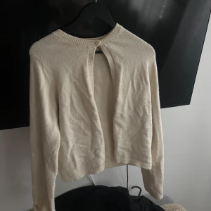 Beige kofta med knappdetalj - Säljer en stilren beige kofta med en knappdetalj vid halsen. Koftan har långa ärmar och är perfekt för lager-på-lager-stil. Passar bra till både vardag och mer uppklädda tillfällen.