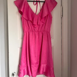 Rosa klänning från Top Secret - Snygg rosa klänning från Top Secret med volangdetaljer och knytband i nacken. Perfekt för sommarens alla tillfällen. Klänningen har en luftig passform och är tillverkad i ett glansigt material som ger en lyxig känsla.