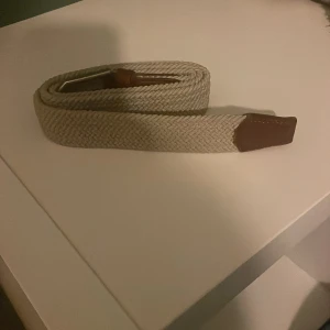 Beige flätat bälte med bruna detaljer - Snyggt beige flätat bälte med bruna skinnändar och silverfärgat spänne. Perfekt för att ge en stilren touch till din outfit. Passar både till jeans och chinos.