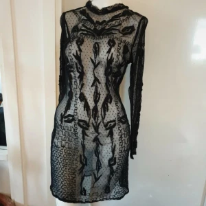 Svart transparent klänning - Snygg svart transparent klänning med långärmad design och detaljerat mönster. Klänningen har en hög hals och knäppning i nacken. Aldrig använd.