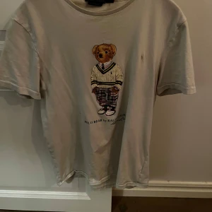 Beige t-shirt med Polo Bear från Ralph Lauren - Säljer en beige t-shirt från Ralph Lauren med ett tryck av den ikoniska Polo Bear i preppy kläder. T-shirten har korta ärmar och rund hals. Perfekt för en avslappnad stil med en touch av klassisk charm. Har en fläck på framsidan annars jättefin!