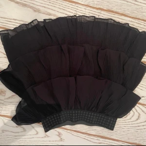 Svart plisserad kjol - Elegant svart plisserad kjol med volangdetaljer. Kjolen har en elastisk midja för bekväm passform och är perfekt för en stilren look. Materialet ger ett fint fall och en luftig känsla. Har inbyggda ”shorts under” aldrig använd