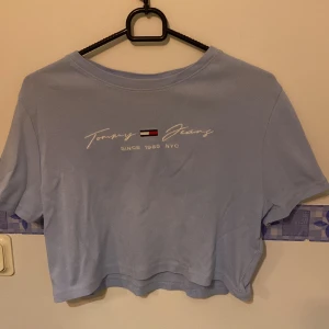 Ljusblå croppad t-shirt från Tommy Jeans - Säljer en ljusblå croppad t-shirt från Tommy Jeans med tryck framtill. Perfekt för en avslappnad stil. T-shirten har korta ärmar och rund halsringning.