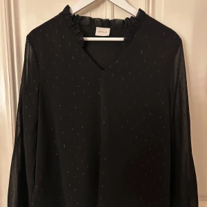 Svart blus från Vila - Elegant svart blus från Vila med subtila prickar och v-ringning. Blusen har långa ärmar och en lätt transparent look som ger en sofistikerad touch. Perfekt för att lyfta vilken outfit som helst.